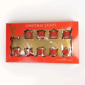 May include: A rectangular red box containing ten drum-shaped Christmas lights. The packaging features the text CHRISTMAS LIGHTS and 110 VOLTS 10 LIGHTS. Each light is red with gold trim and a crisscross pattern, visible through a clear plastic window.