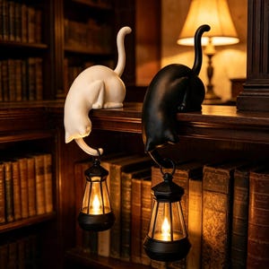 May include: Two cat-shaped ornaments, one white and one black, perched on a ledge. Each cat has a long, upright tail and holds a small, black, lantern-style light fixture suspended from its tail. The lanterns emit a warm, glowing light.