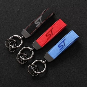 Puede incluir: Tres llaveros con una correa texturizada en negro, rojo y azul. Cada correa tiene las letras ST en relieve en la superficie y está unida a una pieza de metal oscuro con un anillo circular y anillos más pequeños para las llaves.
