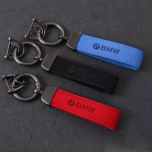 Puede incluir: Tres llaveros con una correa rectangular en azul, negro y rojo, cada uno con un accesorio de metal oscuro y una anilla circular. El logotipo y el texto de BMW están grabados en la superficie de cada correa, que tiene un acabado texturizado.