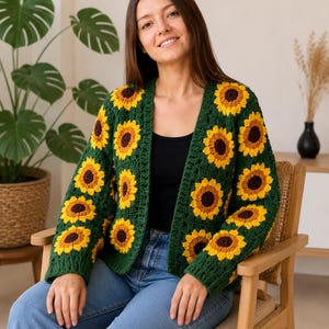May include: A long-sleeved crochet cardigan featuring a repeating pattern of sunflower motifs. The sunflowers have dark brown centers surrounded by bright yellow petals, all set against a deep forest green background. The cardigan has an open front.
