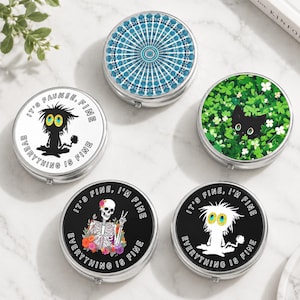 May include: Five round metal pill cases with various designs: one with a blue mandala pattern, one with a black cat in green clover, one with a white skeleton and flowers, and two with a spiky-haired creature and the text IT'S FINE, I'M FINE.
