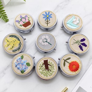 以下が含まれることがあります： ベージュの布地に様々な花の刺繍が施された、8つの丸い銀色のコンパクトミラーのコレクション。ラベンダー、デイジー、アジサイ、ユリなど、色鮮やかな花々がそれぞれのミラーの表面に刺繍されています。