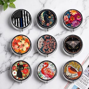 Pode incluir: Nove porta-comprimidos redondos de metal com bordas prateadas, cada um com um design único no topo, incluindo uma bandeira com uma linha azul fina, um gato preto com uma lua crescente, flores coloridas, uma mariposa e um flamingo.