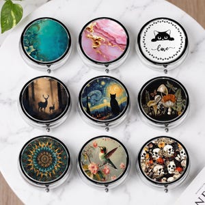 May include: Nine circular metal pill boxes arranged in a grid, each displaying a distinct design: teal floral, pink marble, a cat with the word love, deer in a forest, a cat under a starry sky, mushrooms, a mandala, a hummingbird, and skulls.