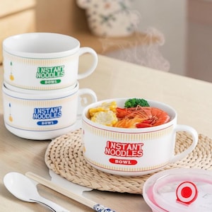 May include: Three white ceramic bowls with handles, each featuring the text "INSTANT NOODLES BOWL" in green, blue, or red. One bowl is filled with shrimp, corn, and broccoli. Includes a white spoon, chopsticks, and a lid with a red circular vent.