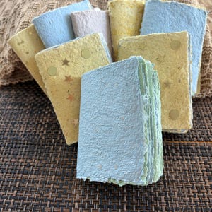 May include: A collection of small, rectangular handmade paper notebooks with deckled edges. The covers feature various pastel colors including light blue, yellow, and beige, with some decorated with gold foil star and circle shapes.