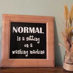Könnte beinhalten: Ein quadratischer Holzrahmen mit hellbraunem Finish enthält ein schwarzes Schild mit weißem Text: NORMAL Is a setting on a washing machine. Der Rahmen steht neben einem durchsichtigen Glasgefäß, das mit getrocknetem Weizen und Gräsern gefüllt ist.