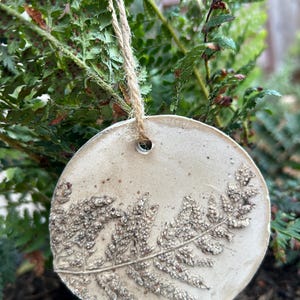 May include: A circular, off-white ceramic ornament featuring a detailed, embossed fern leaf impression on its surface. The ornament has a small hole at the top, threaded with a piece of natural jute twine for hanging, creating a textured botanical design.