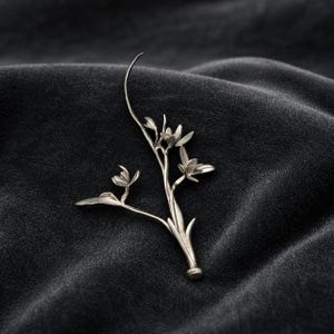 Op de afbeelding: Een zilverkleurige metalen broche in de vorm van een slanke bloeiende tak. Het sieraad heeft drie kleine, delicate bloesems en puntige bladeren die uitsteken vanaf een centrale steel die naar boven toe buigt in een dunne punt.