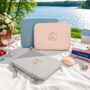 May include: Three rectangular zipped laptop sleeves in light blue, soft pink, and light grey. The blue sleeve features a crown icon and the name Maggie. The pink sleeve displays a mountain graphic and the name Oliver. The grey sleeve shows the name Clara with a bow icon.