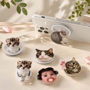 Op de afbeelding: Een collectie ronde telefoonhouders met diverse ontwerpen, waaronder drie verschillende kattengezichten, een kat met een roze bloem en een babygezichtje met een fopspeen. Eén houder is bevestigd aan een transparant telefoonhoesje met magneetring.