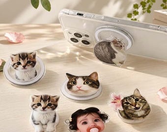 May include: A collection of circular phone grips featuring various designs, including three different cat faces, one cat with a pink flower, and one infant face with a pacifier. One grip is attached to a clear smartphone case with a circular magnetic ring.