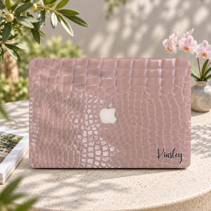 May include: A light pink laptop hard shell case with a textured crocodile skin pattern. The case features a centered cutout for the Apple logo and the name Kinsley printed in black script in the bottom right corner of the cover.
