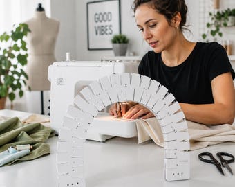 May include: A white, semi-circular wooden darning loom with evenly spaced notches along the outer edge and small holes near the base, positioned on a white surface in front of a sewing machine, fabric, and black scissors.