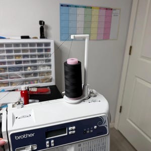 May include: A white Brother sewing machine featuring a digital display, a blue control panel, and a vertical white thread stand holding a large spool of black thread with a pink center. A thin black thread extends from the spool to the machine.