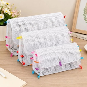 May include: Three white mesh storage pouches of varying sizes, each featuring a grid pattern and secured with colorful plastic safety pins in shades of blue, red, yellow, orange, and purple along the edges and the front flap closure.