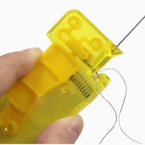 May include: A bright yellow plastic needle threader tool featuring a spring-loaded mechanism. The device has a translucent yellow body with a small metal needle inserted into the top section, with a thin grey thread passing through the eye.