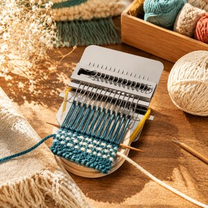 May include: A metal darning loom tool with a circular wooden base, featuring a partially woven patch in teal and white yarn. The device includes a series of vertical metal hooks and a sliding mechanism, with a wooden needle resting on the fabric.