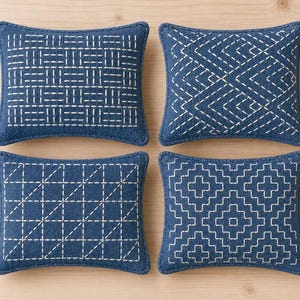 May include: Four rectangular throw pillows in deep blue fabric, each featuring distinct white stitched geometric patterns. The designs include horizontal and vertical dashes, chevron zigzags, diagonal grid lines, and a repeating stepped diamond motif.