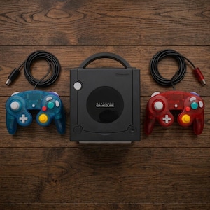Può includere: Una console Nintendo GameCube nera con maniglia superiore e coperchio circolare per il disco, affiancata da due controller cablati. Il controller sinistro è blu traslucido e quello destro è rosso traslucido, entrambi con pulsanti multicolore.