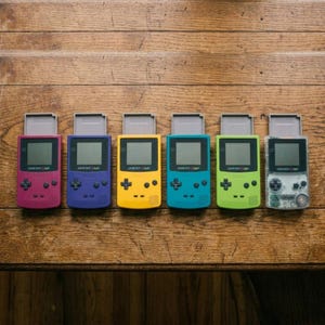 以下が含まれることがあります： 一列に並んだ6台の携帯型ゲーム機「ゲームボーイカラー」。本体カラーはベリー、パープル、イエロー、ティール、ライムグリーン、クリアで、それぞれ十字キー、2つのアクションボタン、画面、そしてグレーのゲームカートリッジが挿入されています。