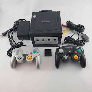 Puede incluir: Una consola Nintendo GameCube negra con panel frontal plateado, un mando con cable plateado y otro negro, un adaptador de corriente negro, un cable AV multicolor y una pequeña tarjeta de memoria negra. Incluye el logo Nintendo GameCube.