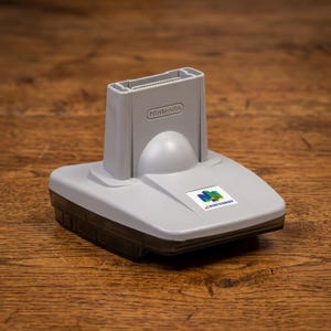 Könnte beinhalten: Ein graues Nintendo 64 Expansion Pak Zubehörteil mit rechteckiger Basis und vertikalem Schlitz. Das Wort Nintendo ist auf der Vorderseite eingeprägt und ein bunter Nintendo 64 Logo-Aufkleber befindet sich auf der Oberseite des Kunststoffgeräts.