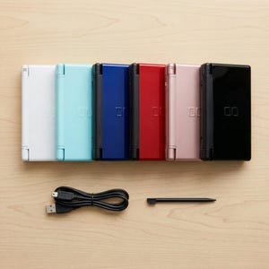 Puede incluir: Seis consolas de juegos portátiles dispuestas en fila, en colores blanco, azul claro, azul oscuro, rojo, rosa y negro. Cada consola tiene una bisagra negra y el logotipo DS. Un cable de carga USB negro y un lápiz óptico negro están debajo.