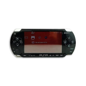 Pode incluir: Uma consola de jogos portátil preta com um ecrã retangular largo que apresenta uma interface de menu. O dispositivo possui um pad direcional à esquerda, quatro botões de ação à direita e os logótipos da Sony e PSP na parte frontal.