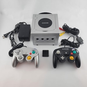 Può includere: Una console Nintendo GameCube color argento con alimentatore nero, due controller in argento e nero, una memory card nera e un cavo AV. La console presenta un coperchio circolare per il disco con il logo Nintendo GameCube stampato in bianco.