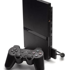 May include: A black Sony PlayStation 2 video game console standing vertically with a matching black wired controller in the foreground. The console features a disc tray, power button, and ports, while the controller includes dual analog sticks.