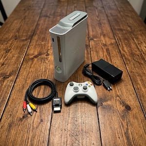May include: A white Xbox 360 game console standing vertically, accompanied by a white wireless controller, a black power brick with a power cord, and a black AV cable with red, white, and yellow RCA connectors for audio and video output.