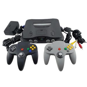 May include: A charcoal grey Nintendo 64 video game console with a black controller, a light grey controller, a black power brick, and a multi-coloured AV cable. The console features a cartridge slot, four controller ports, and the Nintendo 64 logo.