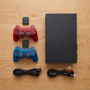 May include: A black rectangular video game console, one translucent red controller, one translucent blue controller, two black memory cards, a black power cord, and a multi-colored AV cable with yellow, white, and red connectors for audio and video.