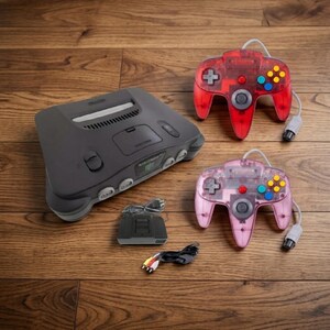 May include: A charcoal grey Nintendo 64 video game console with a cartridge slot, two wired controllers in translucent red and translucent pink, a black expansion pack, and a multi-coloured AV cable with yellow, white, and red connectors.