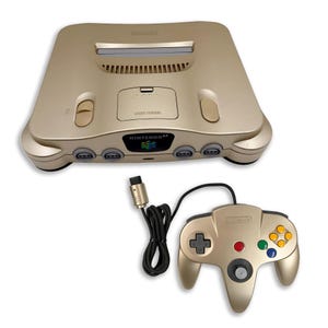 Może przedstawiać: Złota konsola do gier wideo Nintendo 64 z gniazdem na kartridże, czterema portami na kontrolery i przyciskiem zasilania. W zestawie znajduje się pasujący złoty kontroler przewodowy z szarym drążkiem analogowym i czterema kolorowymi przyciskami.