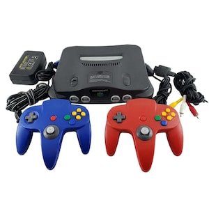 Może przedstawiać: Grafitowa konsola do gier wideo Nintendo 64 z czarnym zasilaczem, niebieskim kontrolerem, czerwonym kontrolerem i kablem AV. Konsola posiada gniazdo na kartridże, cztery porty kontrolerów oraz logo Nintendo 64 na przednim panelu urządzenia.