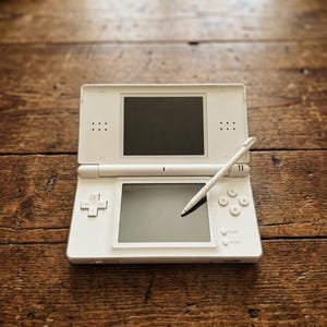May include: A white handheld dual-screen video game console featuring a top display screen, a bottom touchscreen, a directional pad, four action buttons labelled X, Y, A, and B, and Start and Select buttons. A white stylus rests on the bottom screen.