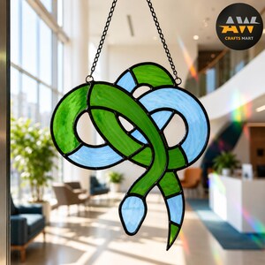 Op de afbeelding: Een glas-in-lood zonnevanger in de vorm van een opgerolde slang, met afwisselende segmenten van doorschijnend groen en lichtblauw glas verbonden door zwarte metalen soldeerlijnen, opgehangen aan een dunne zwarte metalen ketting.