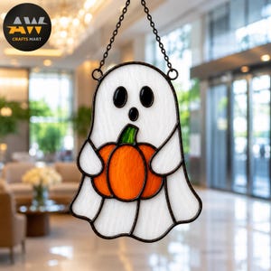 Op de afbeelding: Een glas-in-lood suncatcher in de vorm van een witte geest die een oranje pompoen met een groene steel vasthoudt. De geest heeft twee zwarte ovale ogen en een kleine zwarte ronde mond. Het hangt aan een zwarte metalen ketting.