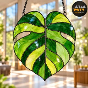 May include: A stained glass suncatcher shaped like a monstera leaf, featuring variegated shades of dark and light green glass segments joined by dark metal solder lines, suspended by a black metal chain with the text AW CRAFTS MART.