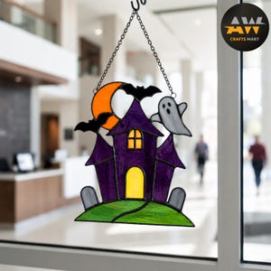 Op de afbeelding: Een glas-in-lood suncatcher met een paars spookhuis met gele ramen, een feloranje halve maan, twee zwarte vleermuizen, een wit spook en twee grijze grafstenen op een groene grasbasis, opgehangen aan een zwarte metalen ketting.