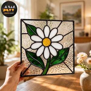 May include: A square stained glass panel featuring a central white daisy with a yellow center and two green leaves. The flower is set against a textured clear glass background, all held together by dark metal lead lines in a geometric frame.