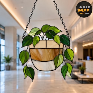 May include: A stained glass suncatcher featuring a potted plant with vibrant green leaves cascading from a beige and white striped pot. The piece is suspended by a black metal chain and includes a circular logo in the corner reading AWL CRAFTS MART.