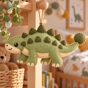 May include: A felt dinosaur ornament in shades of sage green and cream, featuring a row of dark green triangular plates along its back and a round tail tip. The dinosaur has a simple stitched face with a small black eye and mouth.