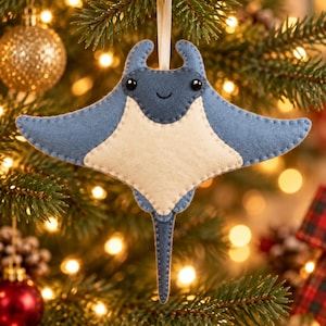 May include: A felt manta ray ornament featuring a blue top and a cream-colored underside. The creature has two small black bead eyes and a stitched smile. A thin ribbon loop is attached to the top for hanging, with visible stitching along the edges.
