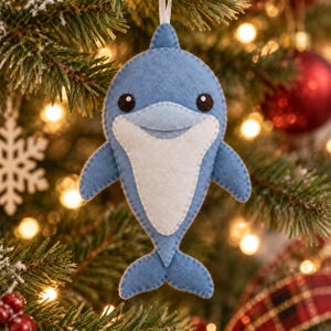 Op de afbeelding: Een dolfijnornament van vilt in blauw- en wittinten. De dolfijn heeft een lichtblauw lichaam met een witte buikvlek, twee zijvinnen, een staart en twee kleine zwarte ronde ogen. Aan de bovenkant is een dun wit lintje bevestigd om het ornament op te hangen.