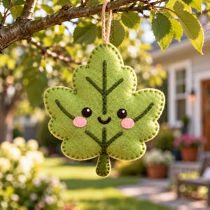 May include: A light green felt leaf ornament featuring a stitched dark green vein pattern, two black eyes, a small smiling mouth, and two pink circular cheeks. The leaf is suspended by a thin brown twine loop from a tree branch.