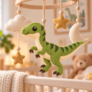 May include: A hanging nursery mobile featuring a central green felt dinosaur with dark green stripes, surrounded by felt shapes including a white cloud, two yellow stars, and a white crescent moon, all suspended from a circular wooden frame.
